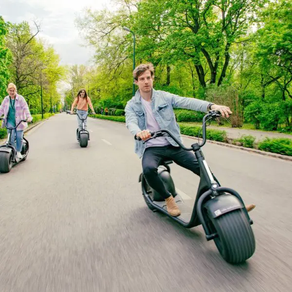 Three people riding electric scooters on a paved road through a green, tree-lined park on a bright day. The leading rider looks at the camera while the two others follow behind, enjoying one of the popular Budapest scooter tours.