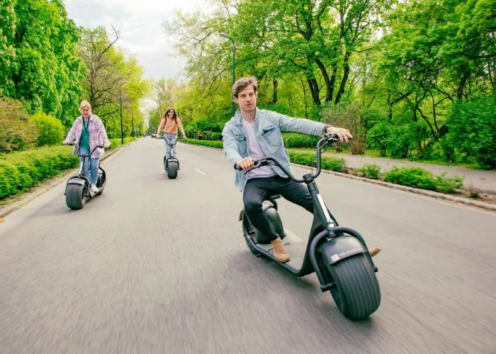 Three people riding electric scooters on a paved road through a green, tree-lined park on a bright day. The leading rider looks at the camera while the two others follow behind, enjoying one of the popular Budapest scooter tours.