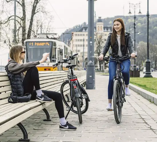 Two women outdoors, one sitting on a bench waving, the other riding a bicycle. Parked bicycles and a tram can be seen in the background along the pathway.