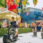 Three people ride electric scooters down a decorated pedestrian street, surrounded by colorful murals and hanging paper lanterns under sunny weather, exploring hidden gems in Budapest.