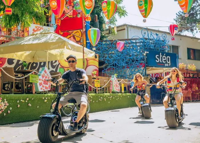 Three people ride electric scooters down a decorated pedestrian street, surrounded by colorful murals and hanging paper lanterns under sunny weather, exploring hidden gems in Budapest.