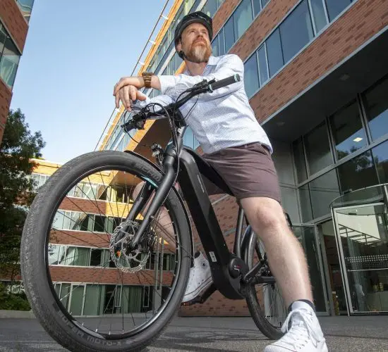 A person with a beard, wearing a helmet, shirt, and shorts, sits on a bicycle in an urban courtyard surrounded by brick buildings and glass windows.