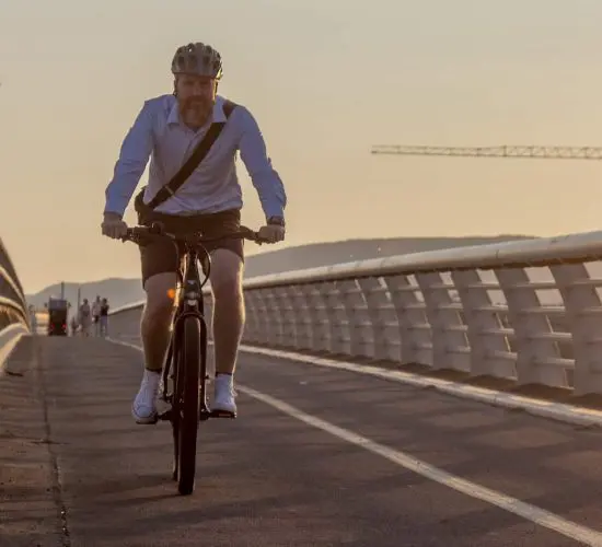 A person wearing a helmet and casual clothing rides a bicycle on a bridge during sunset. Two construction cranes are visible in the background.