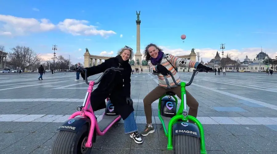 Two smiling people pose on colorful e-scooters in a large open square, with historic monuments and columns in the background under a blue sky—perfect for Budapest sightseeing.