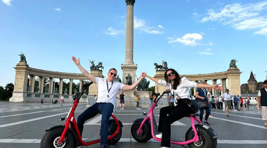 Two people pose on scooters in a large public square with a column and statues in the background. They have their arms outstretched and are smiling.