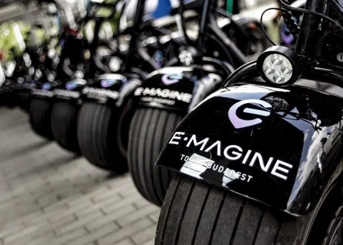 A row of black electric scooters with 'E-MAGINE TOURS BUDAPEST' logos are parked on a tiled ground, showing where and how to rent an electric scooter in Budapest.