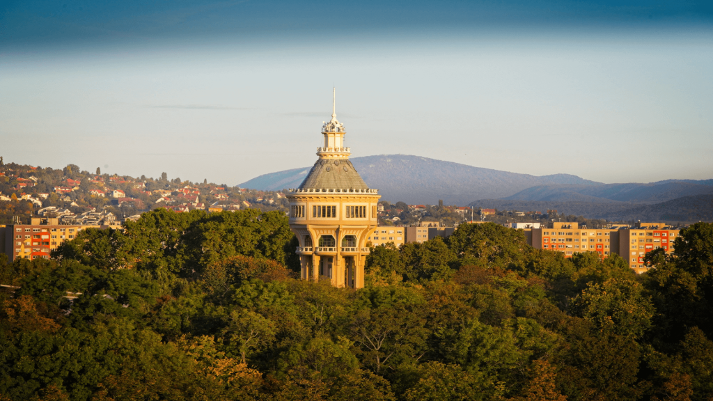 A solitary tower stands in a lush forested area with nearby residential buildings and distant hills under a clear sky, reminiscent of the serene landscapes you might explore when considering things to do in Budapest in autumn.