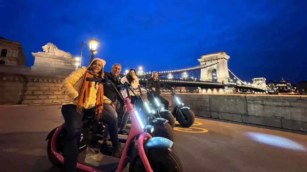 A group of four people ride electric scooters at night near a lit-up suspension bridge.