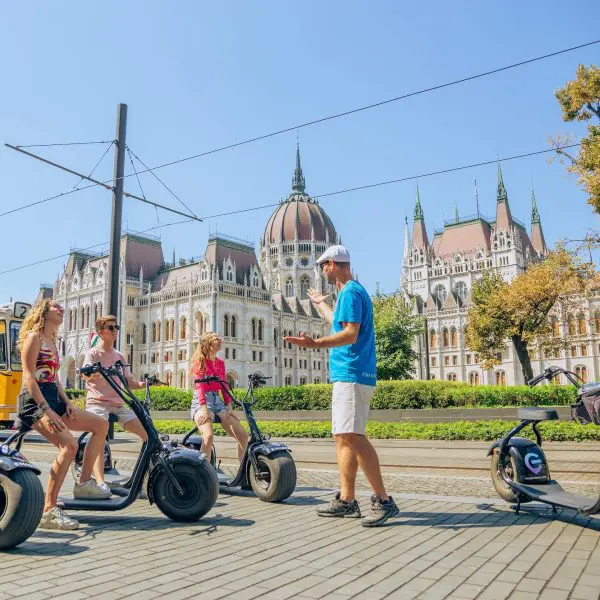 A group of people on electric scooters listens to a guide outside a historic, ornate building on a sunny day.