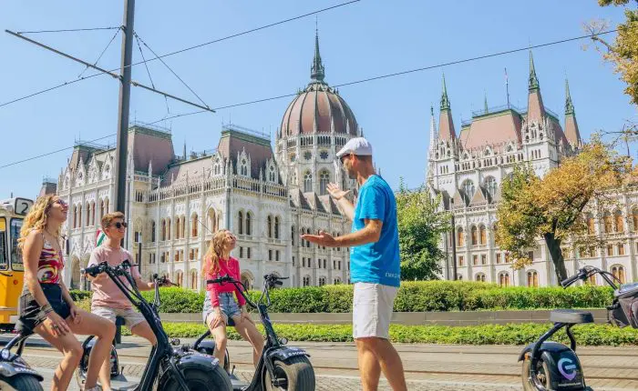 A group of people on electric scooters listens to a guide outside a historic, ornate building on a sunny day.