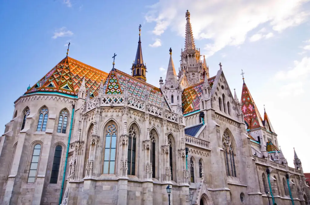 Exterior view of Matthias Church in Budapest, showcasing intricate Gothic architecture and colorful tile roofs under a blue sky with scattered clouds.