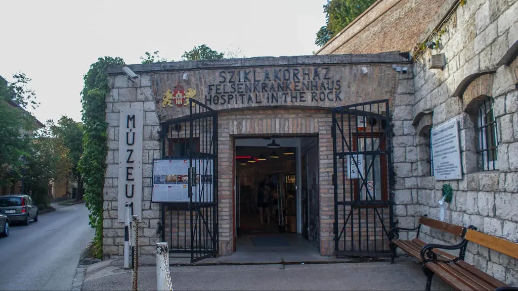 Entrance of the Szlakakorhaz Hospital in the Rock Museum, with an open gate and signage in Hungarian and German. Stone walls surround the entrance, and a street with parked cars is visible on the left.