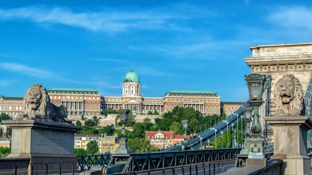 The image shows the Chain Bridge with lion statues in the foreground and Buda Castle in the background under a clear blue sky.