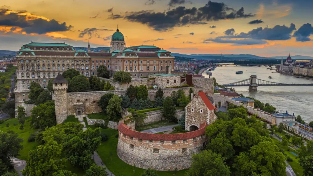 Aerial view of the Buda Castle complex in Budapest, Hungary, at sunset, with the Danube River and cityscape in the background.