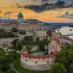 Aerial view of the Buda Castle complex in Budapest, Hungary, at sunset, with the Danube River and cityscape in the background.