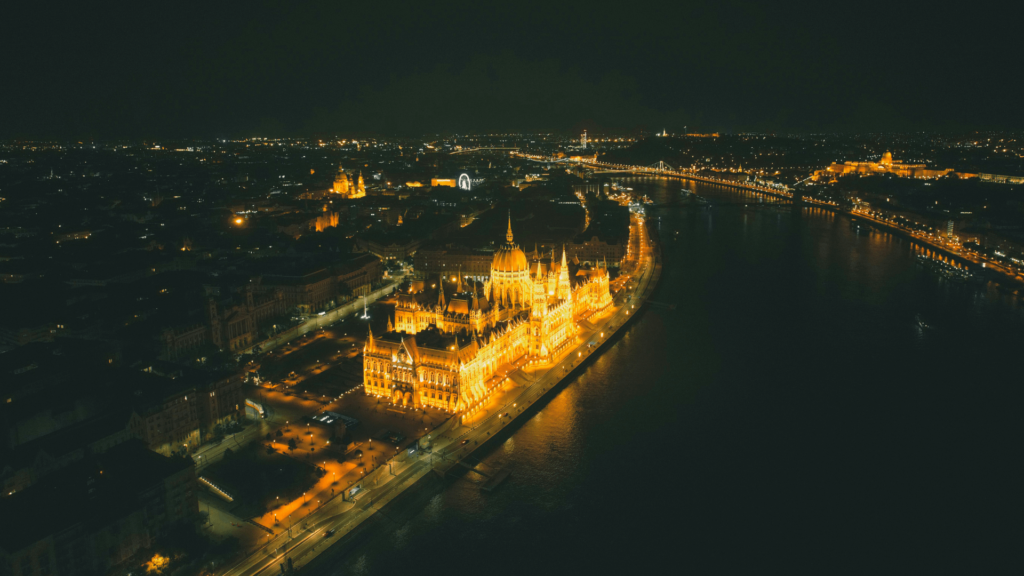 Aerial view of a city at night, featuring a brightly lit building along a river, with lights reflecting on the water and the surrounding cityscape in the background.