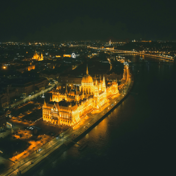 Aerial view of a city at night, featuring a brightly lit building along a river, with lights reflecting on the water and the surrounding cityscape in the background.