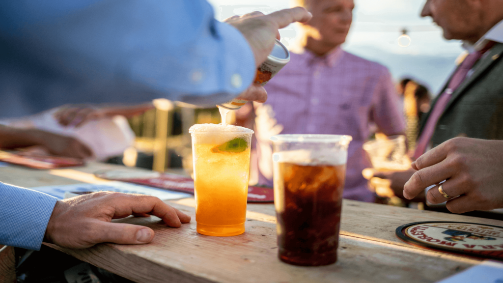 A person pours a beverage into a plastic cup on a wooden counter; another drink sits nearby, with people chatting in the background.