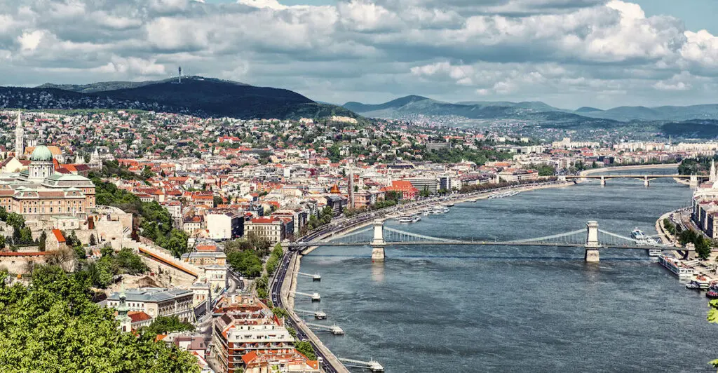 Aerial view of Budapest, showing the Danube River flowing through the city. Bridges connect the Buda and Pest sides, with historic buildings and green hills in the background. Consider an E-Scooter rental to explore this stunning scene on your Budapest trip.