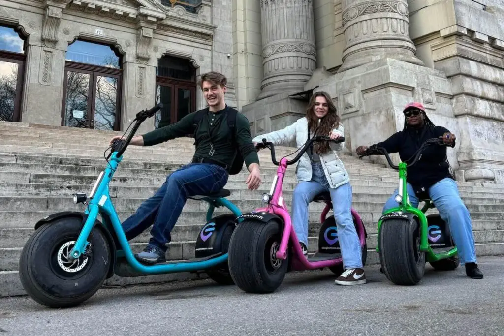 Three people pose cheerfully on rented e-scooters in front of a large, ornate building with stone steps during their Budapest trip. The scooters are bright blue, pink, and green, perfectly matching the groups relaxed vibe.