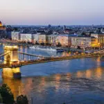 A twilight view of Budapest’s Chain Bridge spanning the Danube River, with city lights reflecting on the water and the Hungarian Parliament building illuminated in the background—a must-see highlight in any Budapest guide.