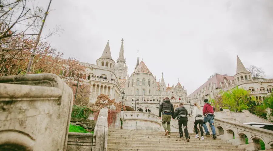 HyperEmagine-43 A group of people climb wide stone steps toward a historic, ornate building with towers and a pointed roof under a cloudy sky, surrounded by trees and greenery.