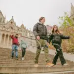 A group of young adults walk down wide stone steps in front of an ornate historic building with towers on a cloudy day. Two people smile in the foreground, enjoying their Buda Castle tour in Budapest while others follow behind.