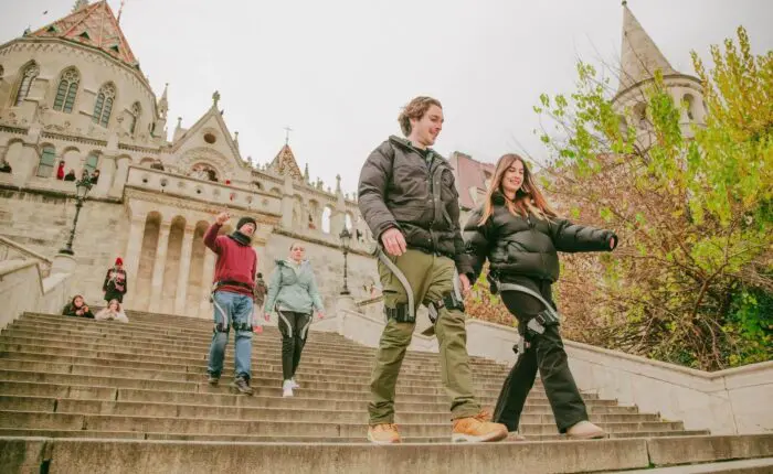 A group of young adults walk down wide stone steps in front of an ornate historic building with towers on a cloudy day. Two people smile in the foreground, enjoying their Buda Castle tour in Budapest while others follow behind.