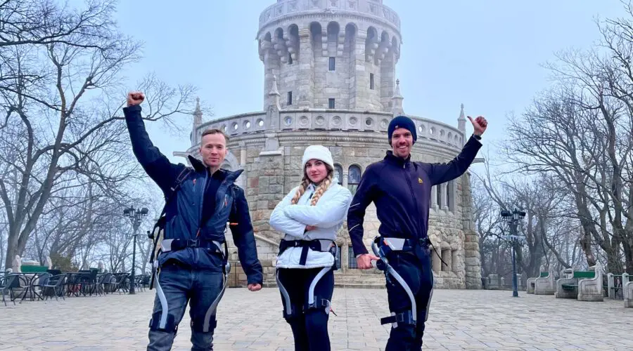 IMG_0261 Three people wearing outdoor gear pose confidently with raised fists in front of a tall, round stone tower on a misty day. Leafless trees and cobblestone pavement surround the scene.