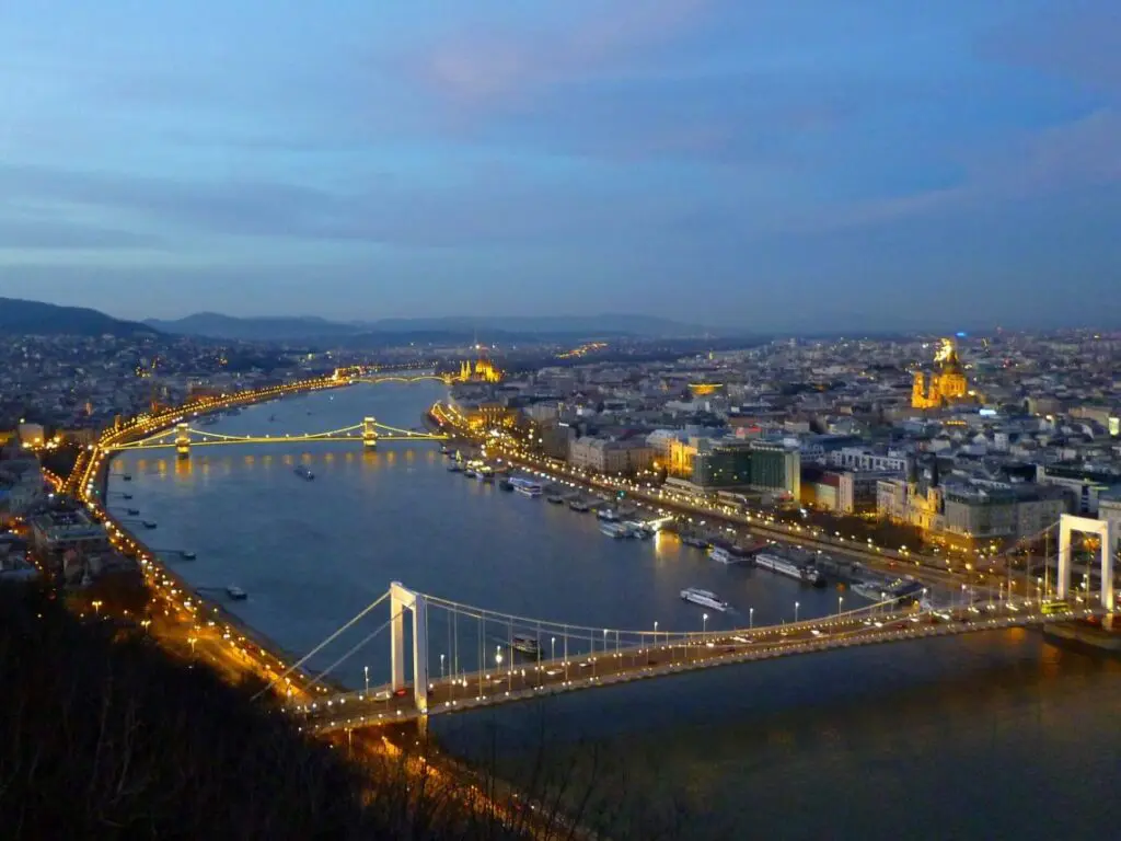 A panoramic evening view of Budapest, Hungary, featuring the Danube River, illuminated bridges, city lights, and the historic Buda Castle District under a dusky blue sky.
