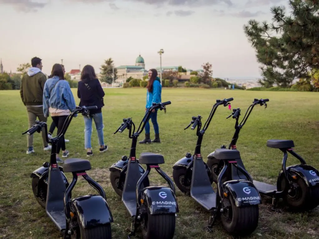 Four people walk on a grassy field toward a distant building in the Buda Castle District, while six parked black electric scooters with E-MAGINE branding are in the foreground. Trees and a cloudy sky complete the Budapest backdrop.
