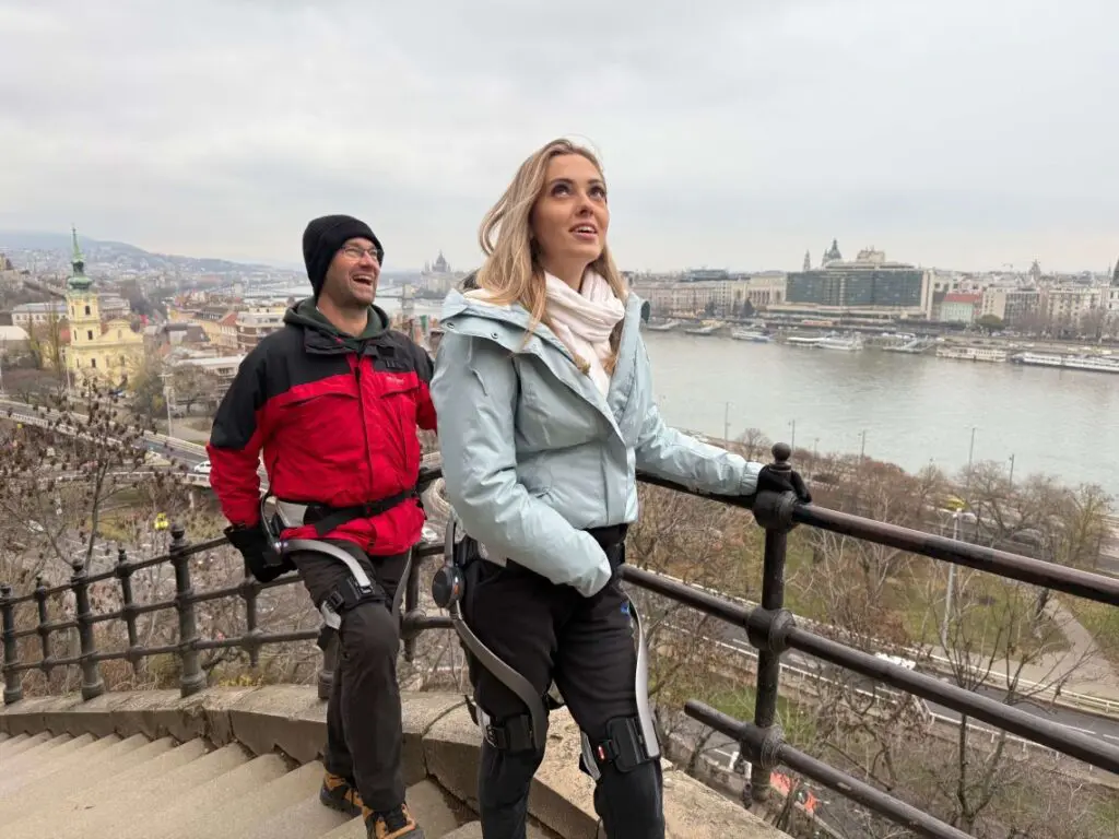 A woman and a man in hiking gear walk up outdoor stairs beside a metal railing, overlooking the river and cityscape near the Buda Castle District on a cloudy day. The man is smiling, while the woman looks ahead with a focused expression.