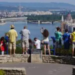 citadel-blog-hero A group of tourists stand on a stone overlook on Gellert Hill Budapest, taking photos of a wide river and cityscape with bridges and historic buildings, including a domed parliament building, under a clear blue sky.