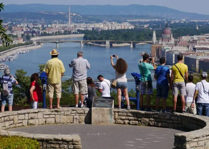 citadel-blog-hero A group of tourists stand on a stone overlook on Gellert Hill Budapest, taking photos of a wide river and cityscape with bridges and historic buildings, including a domed parliament building, under a clear blue sky.