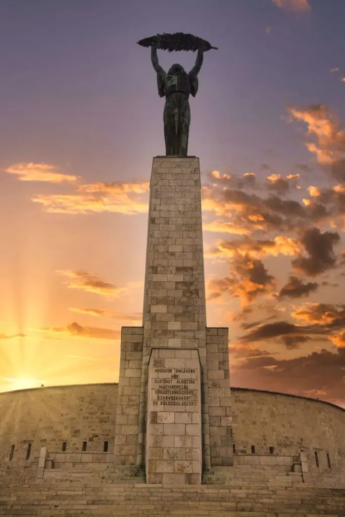 A tall stone monument stands in the Buda Castle District, its statue of a woman holding a palm leaf silhouetted against a dramatic sunset sky with orange clouds. The statue rises atop a pedestal inscribed at the base.