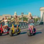 Three people ride colorful electric scooters on a sunny bridge, embodying the spirit of The Happy Hour, with a historic building and statues visible in the background. The mood is cheerful and relaxed.
