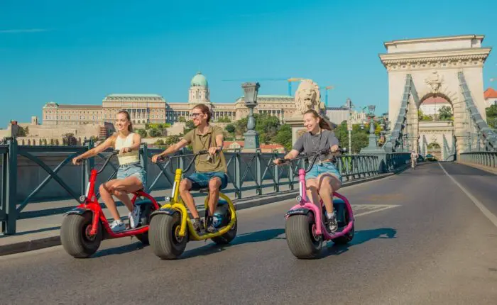 Three people ride colorful electric scooters on a sunny bridge, embodying the spirit of The Happy Hour, with a historic building and statues visible in the background. The mood is cheerful and relaxed.