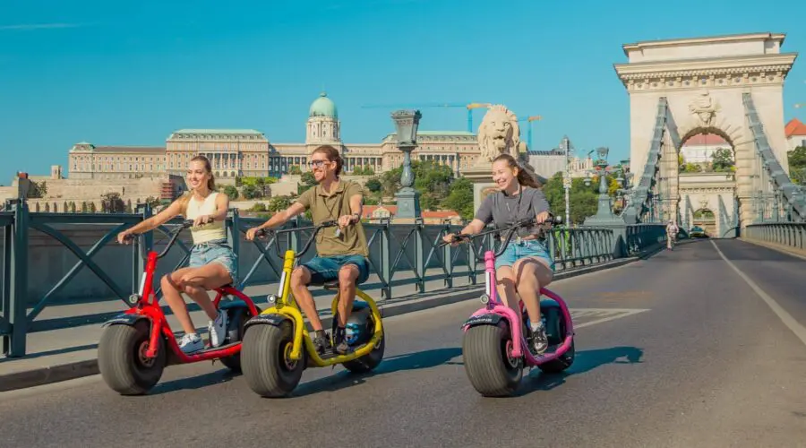 The Happy Hour tour Three people ride colorful electric scooters on a sunny bridge, embodying the spirit of The Happy Hour, with a historic building and statues visible in the background. The mood is cheerful and relaxed.