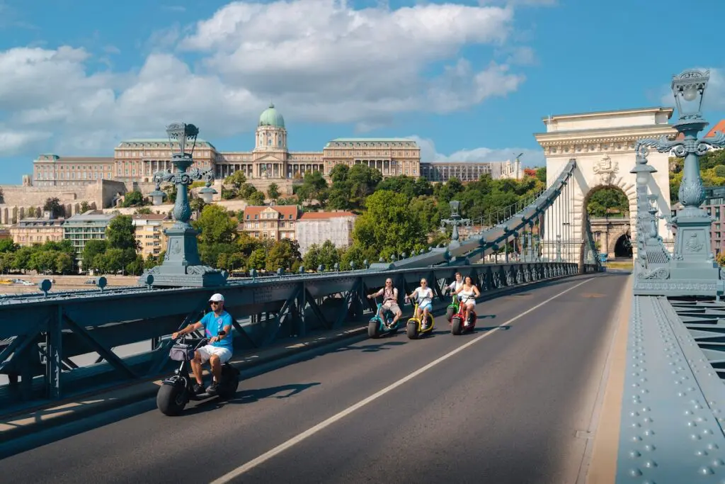 People ride e-scooters on the Chain Bridge in Budapest with a historic cityscape, including a domed building and other classical architecture, visible in the background under a blue sky with clouds.