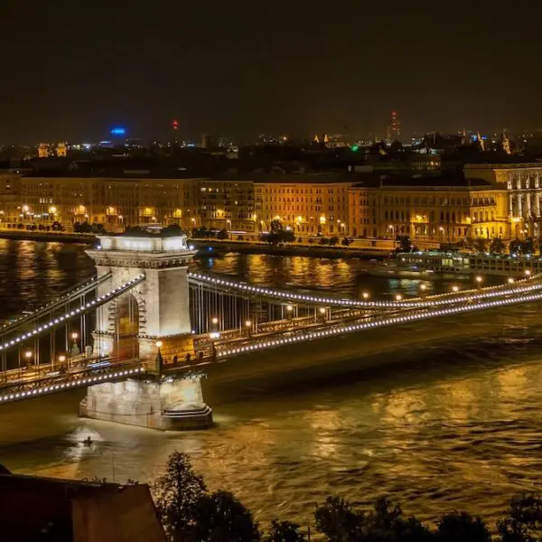 The Chain Bridge in Budapest, Hungary, illuminated at night, spans the Danube River with city buildings and the Hungarian Parliament visible in the background.