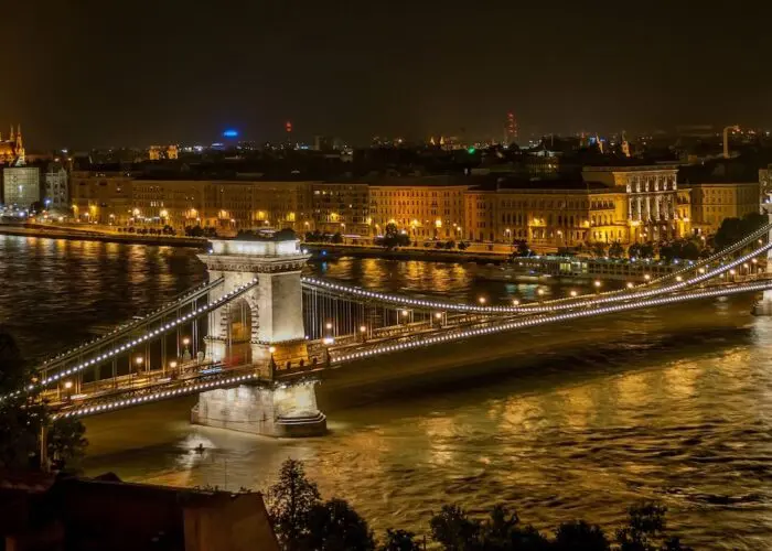 The Chain Bridge in Budapest, Hungary, illuminated at night, spans the Danube River with city buildings and the Hungarian Parliament visible in the background.