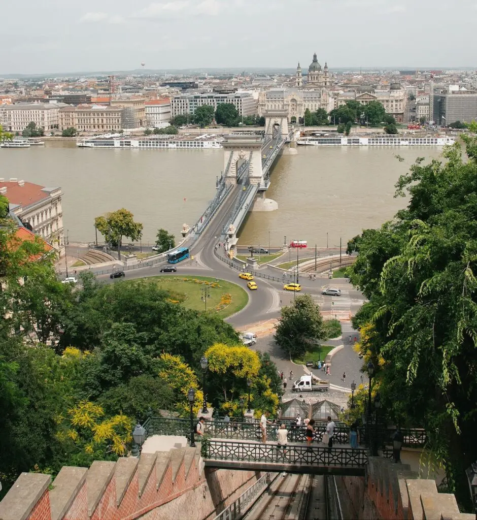 View of Chain Bridge over the Danube River in Budapest, with cars crossing, surrounded by city buildings, greenery, and people riding a funicular in the foreground.