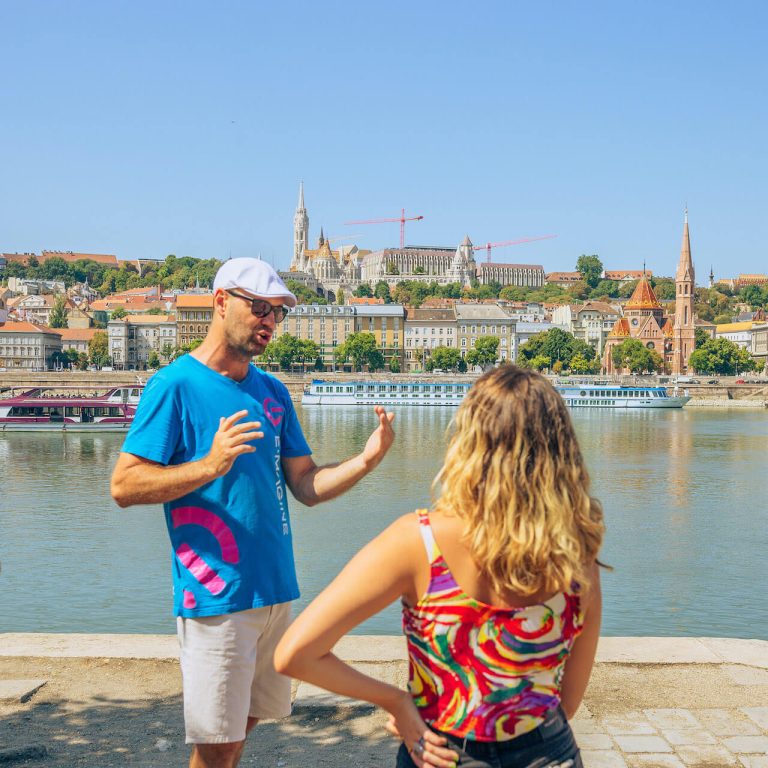 A man wearing a blue shirt and white cap is talking to a woman with blonde curly hair by a river with a city skyline and a docked boat in the background on a sunny day.