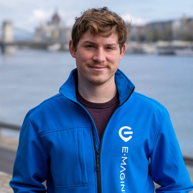 A person with short brown hair wearing a blue jacket with the E-MAGINE logo stands outdoors near a body of water with a cityscape blurred in the background.