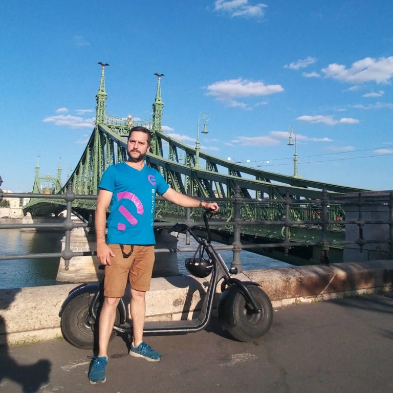 A man wearing a blue T-shirt and brown shorts stands with an electric scooter in front of a green bridge spanning a river, under a clear blue sky.