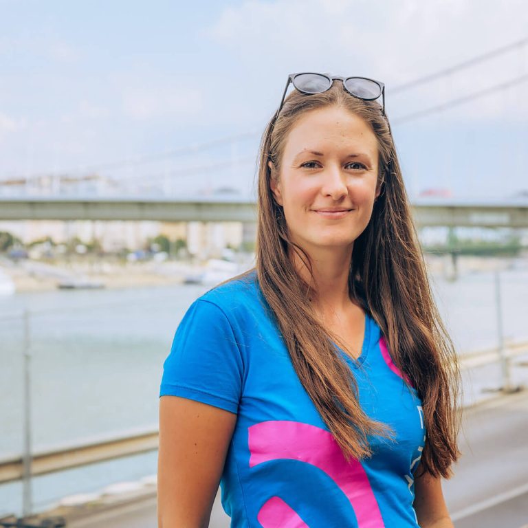 A woman with long brown hair stands outdoors wearing a blue shirt with a pink design and sunglasses on her head, with a bridge and body of water in the background.