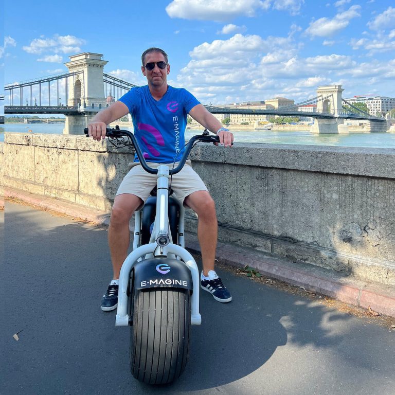 A man in sunglasses sits on an eMagine bike on a paved pathway with a stone balustrade bridge and river in the background.