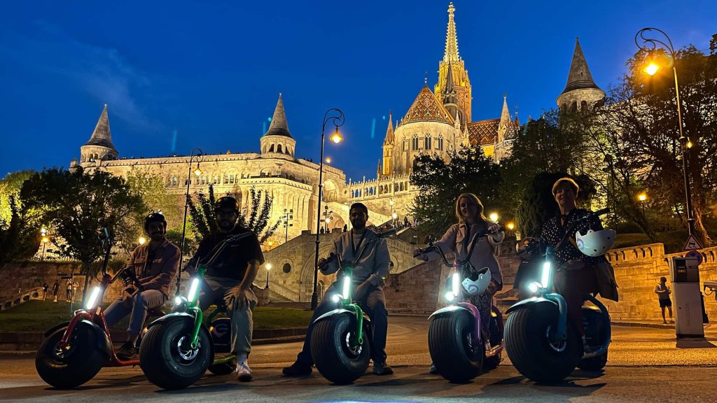 Five people on electric unicycles pose in front of an illuminated building with spires and towers at dusk.