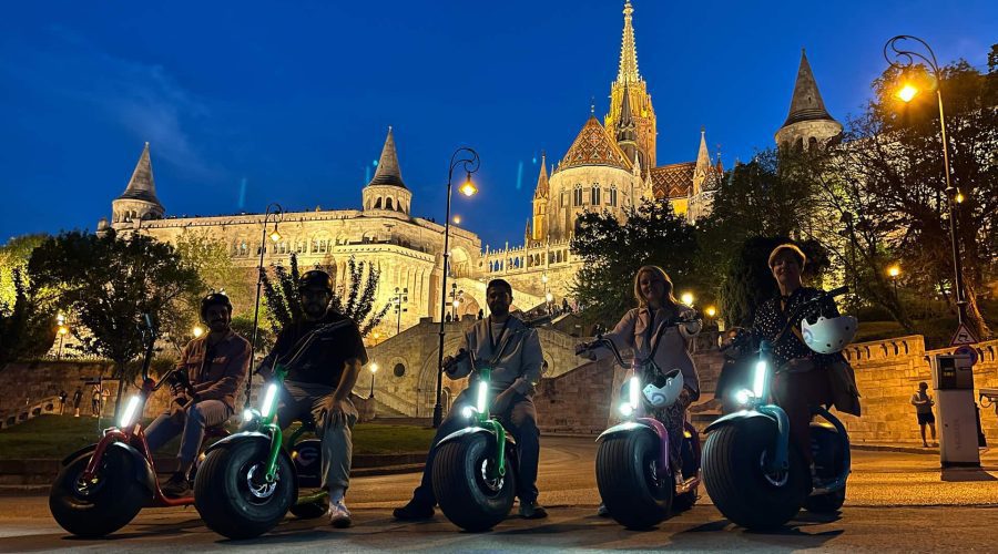 Five people on electric unicycles pose in front of an illuminated building with spires and towers at dusk. Egy ötfős csoport elektromos robogón parkol éjszaka egy kivilágított történelmi épület előtt.