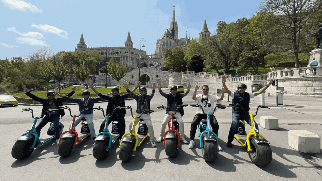 A group of eight people on motorized scooters raise their arms in celebration. They are in an urban park with historic buildings and greenery in the background.